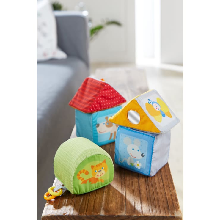 Drei bunte Babyspielzeuge aus weichem Stoff in Form von Häusern und einem Zylinder liegen auf einem Holztisch. Auf den Spielzeugen sind Tiere abgebildet, darunter ein Hund, eine Maus, ein Schmetterling und eine Katze. Im Hintergrund sind ein Sofa und eine Pflanze zu sehen.