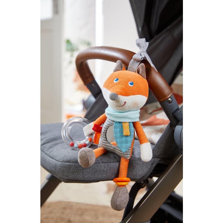 Ein Plüschfuchs mit Schal und Ringrassel ist am Griff eines grauen Kinderwagens befestigt. Das Spielzeug ist orange, weiß und grau, mit blauen Details und einem freundlichen Ausdruck.