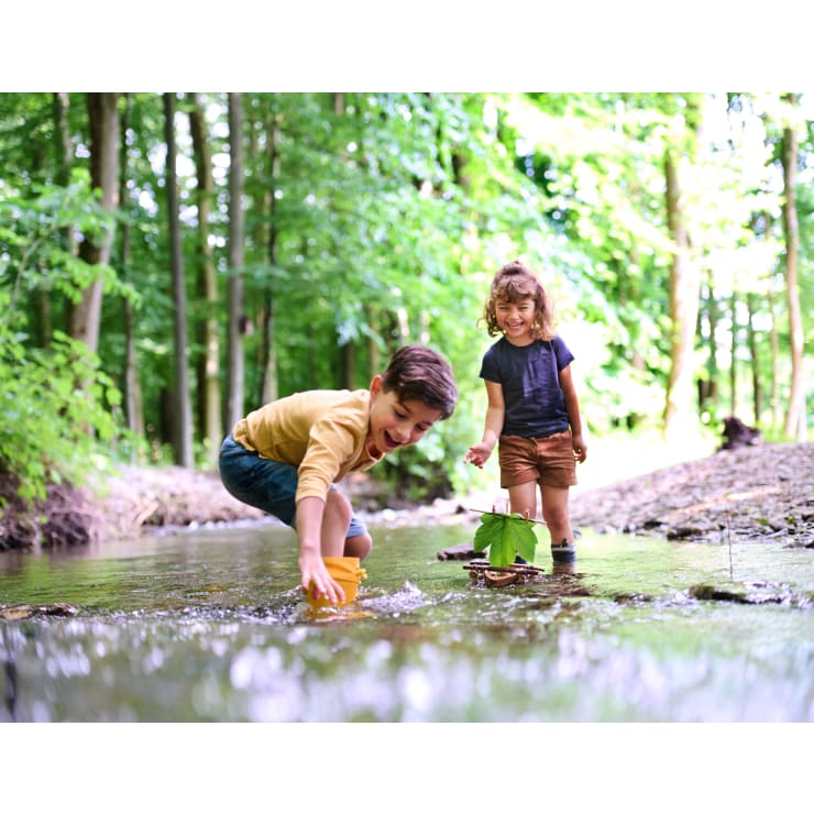 Zwei Kinder spielen in einem seichten, von Bäumen umgebenen Bach. Ein Kind kniet und greift mit einem gelben Behälter ins Wasser, während das andere lächelnd daneben steht. Die Szene ist hell und im Freien.