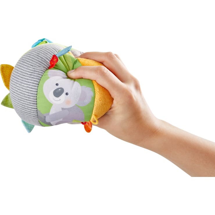 Eine Hand hält ein weiches, farbenfrohes Babyspielzeug mit einer lächelnden Koala-Illustration, verschiedenen Stofftexturen und kleinen Stoffanhängern vor einem weißen Hintergrund.