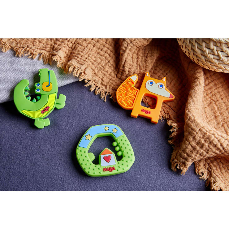 Drei farbenfrohe HABA-Beißringe in Form eines grünen Krokodils, eines orangefarbenen Fuchses und eines grünen Rings mit Hausmotiv liegen auf einer lilafarbenen Fläche neben einer strukturierten braunen Decke und einem geflochtenen Korb.