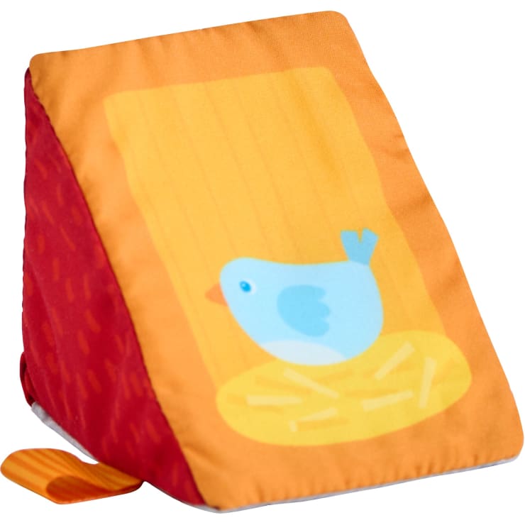 Ein weicher, keilförmiger Stoffblock mit einer roten Seite und einer orangefarbenen Vorderseite, auf der ein blauer Vogel auf einem gelben Nest oder Stroh sitzt.