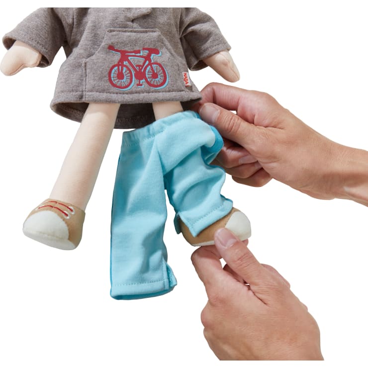 Eine Person kleidet eine Stoffpuppe mit hellblauen Hosen ein. Die Puppe trägt ein graues Oberteil mit einer roten Fahrradgrafik und beigefarbene Schuhe. Das Gesicht der Puppe ist nicht sichtbar.