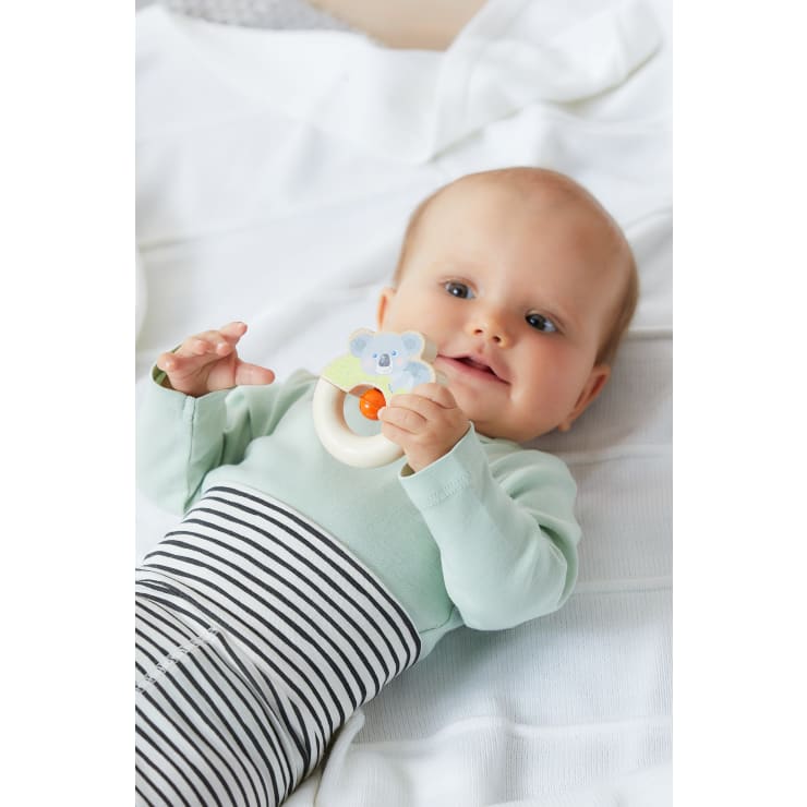 Ein Baby liegt auf einer weißen Decke, trägt ein hellgrünes Oberteil und eine schwarz-weiß gestreifte Hose und hält ein Spielzeug mit einem Koalagesicht und einem orangefarbenen Ring.