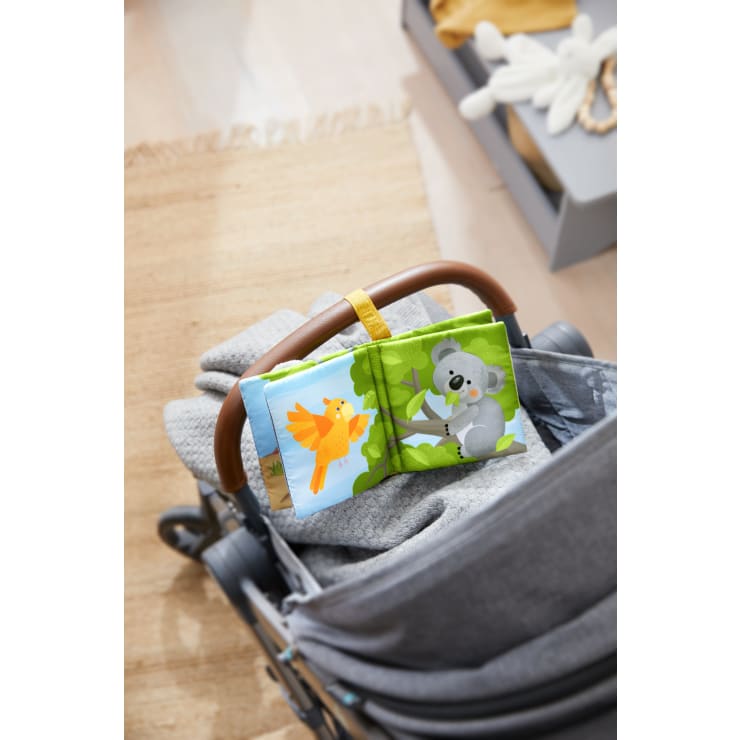 Ein weiches Stoffbuch mit Abbildungen eines Vogels und eines Koalas ist an einem braunen Kinderwagengriff befestigt. Der Kinderwagen ist mit einer grauen Decke bedeckt. Im Hintergrund ist eine Kiste mit Spielzeug zu sehen.