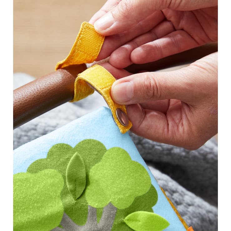 Nahaufnahme von Händen, die ein Stoffband mit einem Klettverschluss und einem Druckknopf um einen Holzstab befestigen. Das Band ist an einem Stofftier mit einem grünen Baummuster auf hellblauem Stoff befestigt.