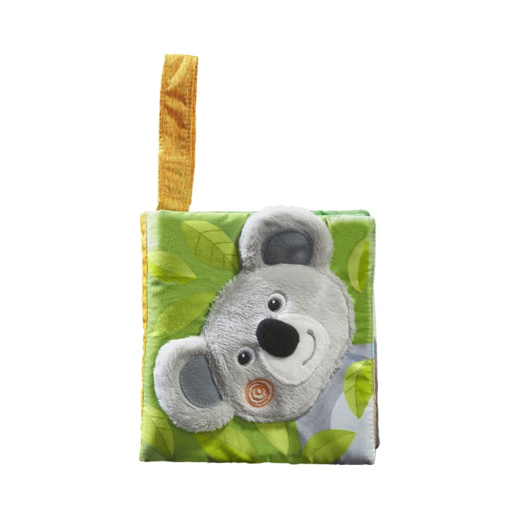Ein kleines Buch aus weichem Stoff mit einem grünen Blattmuster und einem grauen Plüsch-Koala-Gesicht auf dem Einband; eine orangefarbene Stoffschleife ist an der oberen linken Ecke befestigt.