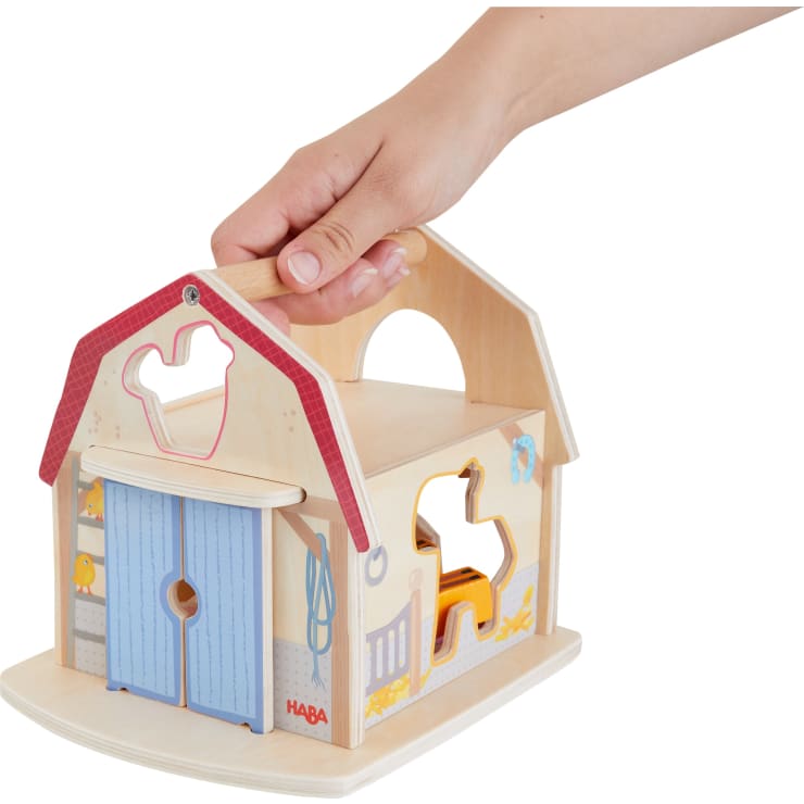Eine Hand hält eine hölzerne Spielzeugscheune mit einem roten Dach, blauen Türen und tierförmigen Ausschnitten an den Seiten. Die Scheune hat oben einen Griff zum Tragen und ist mit bunten Illustrationen und Formen versehen.