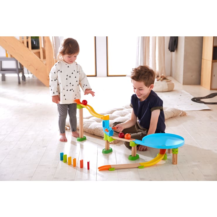 Zwei kleine Kinder spielen mit einer bunten Kugelbahn auf einem hellen Holzboden. Die Bahn verfügt über Rampen, eine Spirale und Dominosteine, die am Ende aufgestellt sind. Im Hintergrund sind Kissen und Holzmöbel zu sehen.