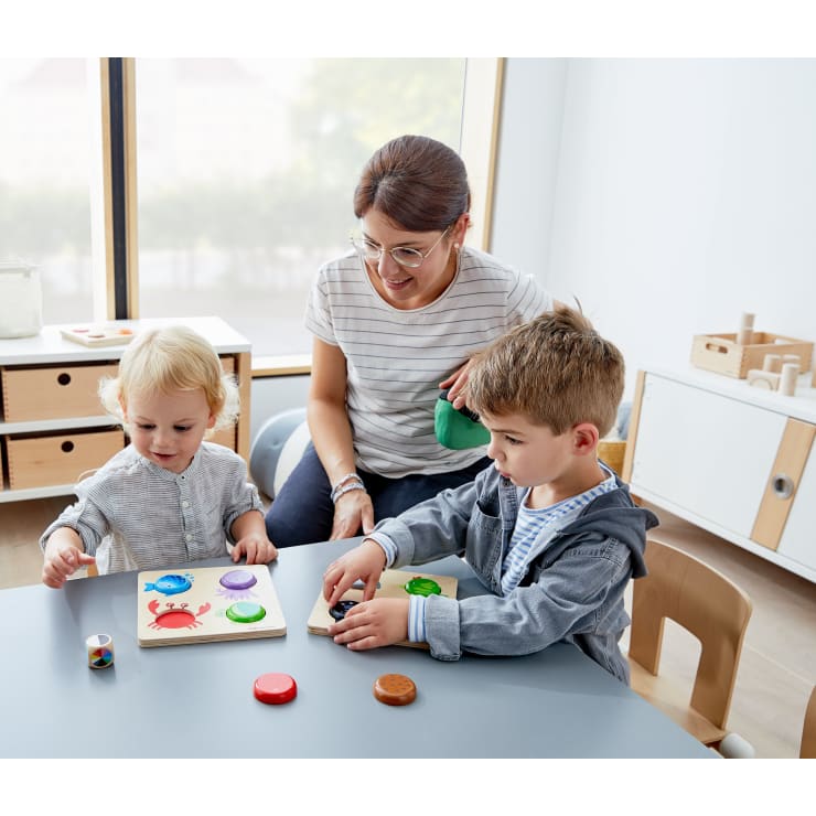 Eine erwachsene Frau beobachtet zwei kleine Kinder, die an einem Tisch sitzen und in einem hellen, aufgeräumten Raum mit Puzzles und bunten Spielfiguren spielen.