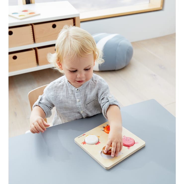 Ein kleines Kind mit blondem Haar sitzt an einem grauen Tisch und legt ein hölzernes Puzzlestück auf ein Puzzlebrett. Im Hintergrund sind ein weiches blaues Kissen und hölzerne Aufbewahrungsschubladen zu sehen.