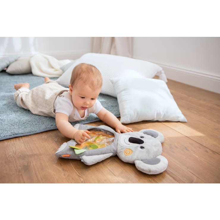 Ein Baby liegt auf einem Holzboden und greift nach einem sensorischen Spielzeug in Form eines Koalas. Weiche Kissen und ein blauer Teppich liegen im Hintergrund, und natürliches Licht erfüllt den Raum.