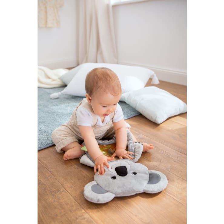 Ein Baby sitzt auf einem Holzboden und spielt mit einem grauen Plüschkoala. Im Hintergrund befinden sich Kissen und ein blauer Teppich in einem hellen, sanft beleuchteten Raum.