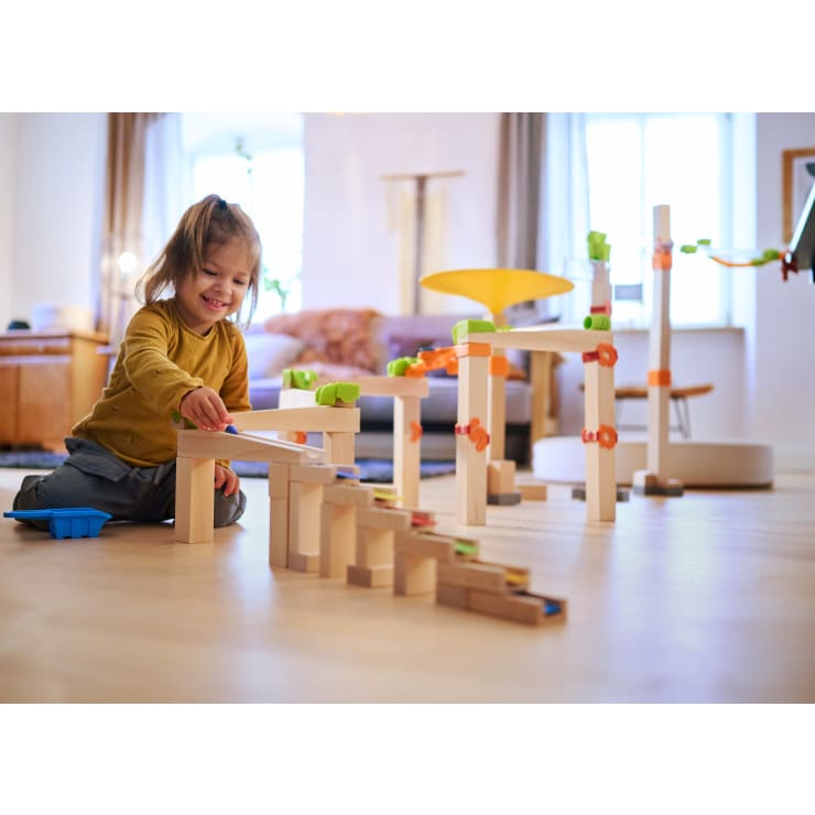 Ein kleines Kind sitzt auf einem Holzfußboden in einem Haus und spielt mit einer Murmelbahn aus Holzklötzen und bunten Plastikteilen. Das Sonnenlicht fällt durch große Fenster im Hintergrund.