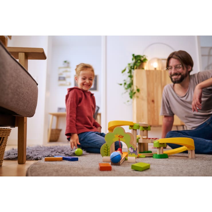 Ein Kind und ein Erwachsener sitzen auf dem Boden und spielen mit einem bunten Holzspielzeug in einem warm beleuchteten Wohnzimmer. Das Kind hält lächelnd einen grünen Ball in der Hand, während der Erwachsene in der Nähe zusieht.