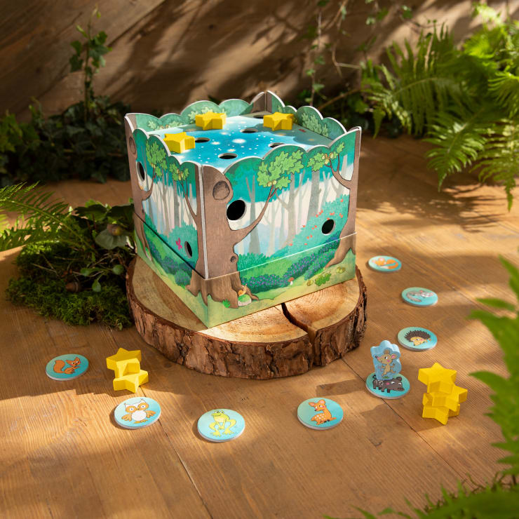 Eine farbenfrohe Kinderspielschachtel mit einem Waldmotiv sitzt auf einem Baumstumpf, umgeben von gelben sternförmigen Spielsteinen und kreisförmigen Tierplättchen, mit Pflanzen und Holzfußboden im Hintergrund.