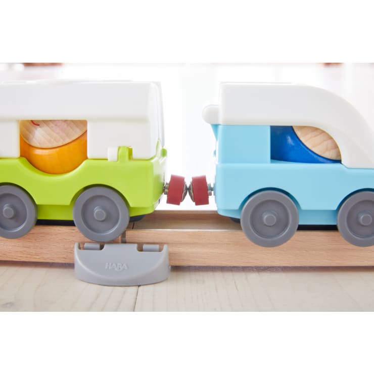 Nahaufnahme von zwei Spielzeugeisenbahnwagen, einem grün-weißen und einem blau-weißen, die auf einer Holzschiene verbunden sind. In jedem Wagen befindet sich eine sichtbare Holzkugel. Die Schiene liegt auf einem hellen Boden.