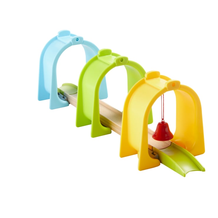 Eine hölzerne Spielzeugeisenbahnstrecke führt durch drei bunte Kunststoffbögen - blau, grün und gelb - und hat eine kleine rote Glocke, die am gelben Bogen hängt.