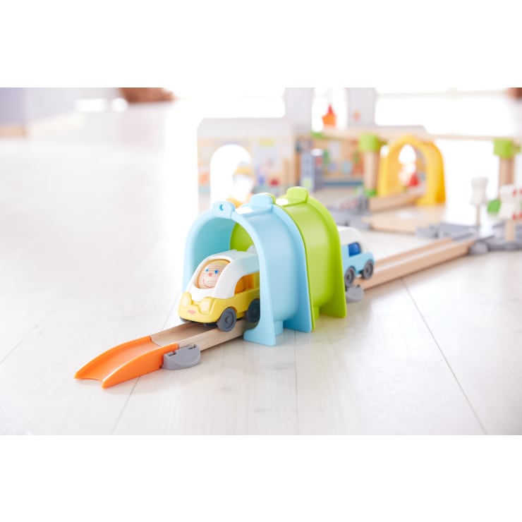 Spielzeugautos aus Holz fahren auf einer Schiene durch blaue und grüne Kunststofftunnel, an deren Ende eine orangefarbene Rampe steht. Die Anlage ist auf einem hellen Holzboden aufgebaut. Im Hintergrund sind weitere Spielzeugkonstruktionen zu sehen.