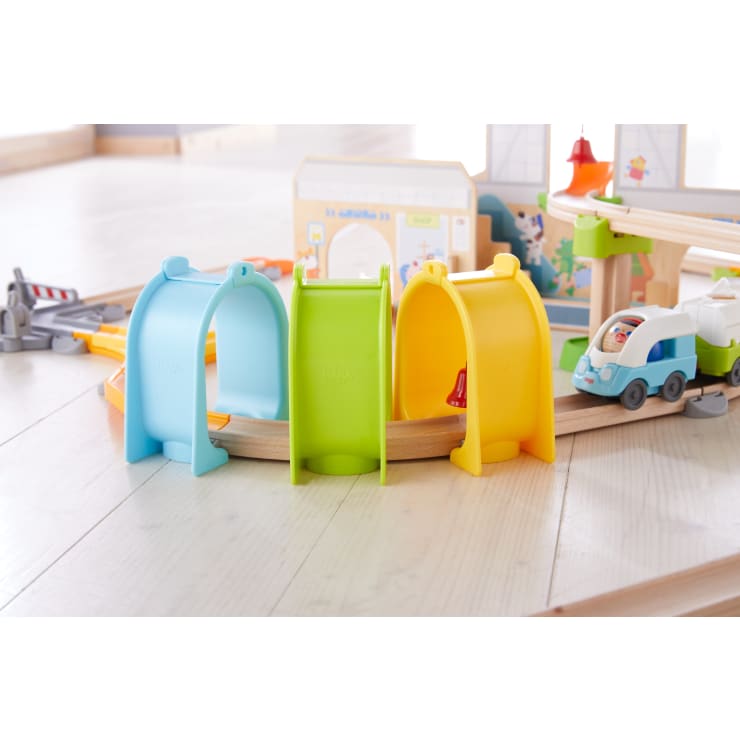 Eine hölzerne Spielzeugeisenbahn steht auf einem hellen Boden, auf dem im Vordergrund drei Plastiktunnel in den Farben Blau, Grün und Gelb aufgereiht sind. Auf den Gleisen ist eine kleine Spielzeugeisenbahn mit einem Lokführer zu sehen.