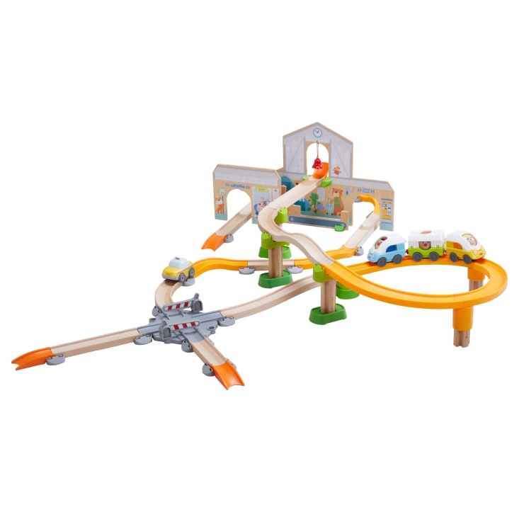 Ein farbenfrohes Spielzeugeisenbahn-Set mit Holz- und Kunststoffgleisen, einer Bahnhofskulisse, einer kleinen Brücke, Verkehrsschranken und einem weißen Spielzeugeisenbahnzug mit Tiergesichtern auf den Wagen. Einige Gleise sind mit grünen Stützen erhöht.