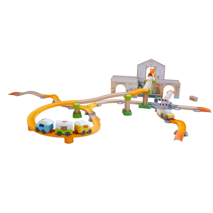 Ein Spielzeugeisenbahnset aus Holz mit bunten Gleisen, Brücken und Rampen, mit einem Zug mit drei Wagen, einem Gebäude mit Bogeneingang und kleinen Spielzeugautos auf der rechten Seite.