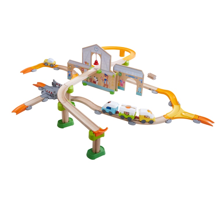 Ein Spielzeugeisenbahnset aus Holz mit bunten Gleisen, erhöhten Abschnitten, einem Bahnhofsgebäude, drei Waggons, einem Taxi, Bäumen und verschiedenen dekorativen Details in einer spielerischen Anordnung.