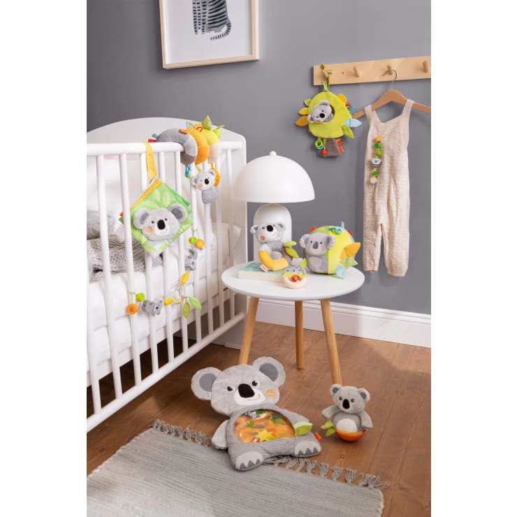 Ein Kinderzimmer mit einem weißen Kinderbett, grauen Wänden, einem runden weißen Tisch, einer Lampe, Koala-Plüschtieren, einem hängenden Strampler und verschiedenen Stofftieren auf dem Kinderbett, dem Tisch und dem Boden.