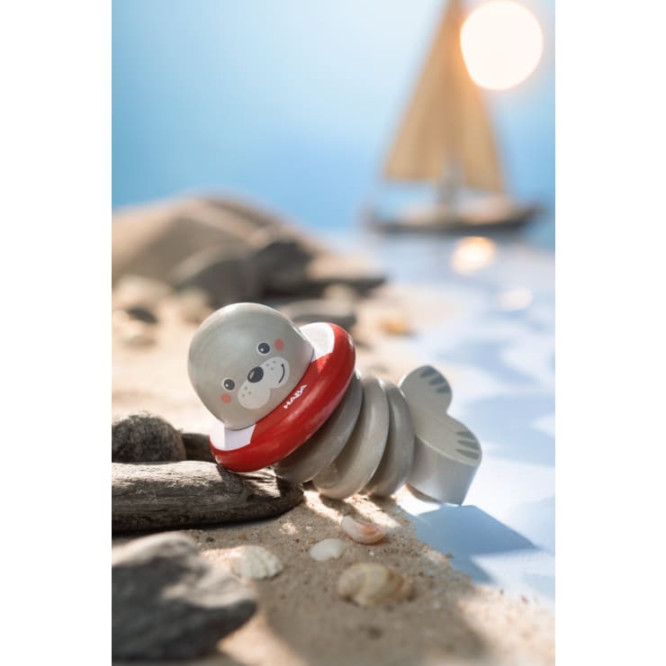 Ein hölzernes Robbenspielzeug mit einer rot-weißen Rettungsboje ruht auf sandigem Grund zwischen kleinen Felsen und Muscheln. Im Hintergrund sind ein verschwommenes Segelboot und Sonnenlicht zu sehen.