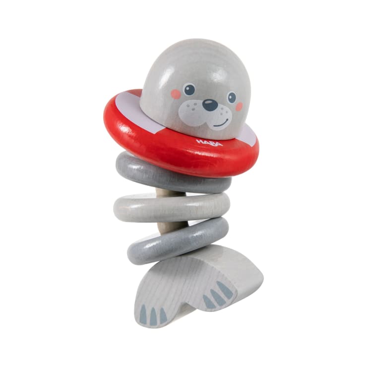 Ein Holzspielzeug in Form einer grauen Robbe mit einem rot-weißen Ring um den Hals, einem gewundenen Federkörper und flachen Flossen. Die Robbe hat ein lächelndes Gesicht und ist vor einem weißen Hintergrund zu sehen.