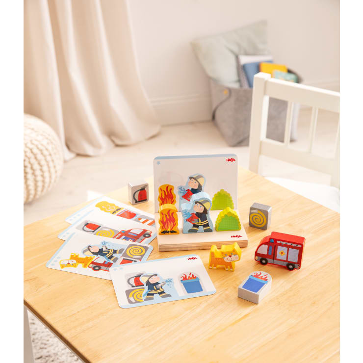 Auf einem Holztisch liegen illustrierte Aktivitätskarten mit Feuerwehrleuten, Spielzeugblöcken, einem kleinen Spielzeug-Feuerwehrauto und einer gelben Hundefigur. Im Hintergrund stehen ein Stuhl, ein weicher Hocker und ein Aufbewahrungskorb mit Büchern.