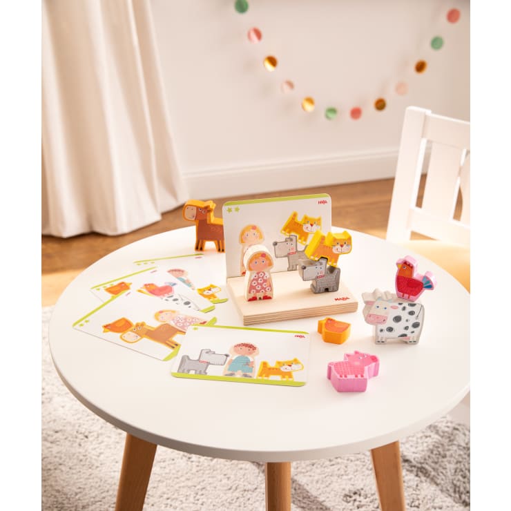 Ein runder weißer Tisch mit Tier- und Personenpuzzleteilen, mehreren illustrierten Karten und einem kleinen Holzständer, der eine Karte und zusammengesetzte Teile zeigt. Im Hintergrund stehen ein weißer Stuhl und bunte Girlanden.