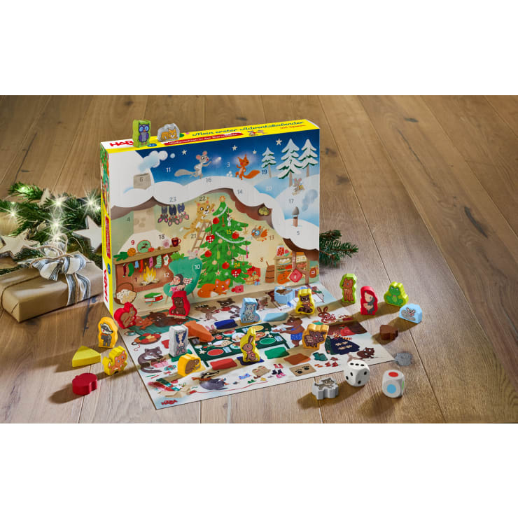 Ein bunter Adventskalender mit illustrierten Tierfiguren, einem geschmückten Weihnachtsbaum und nummerierten Türchen. Davor sind hölzerne Tierspielfiguren, Würfel und ein Spielbrett angeordnet. In der Nähe befinden sich Geschenke und Weihnachtslichter.