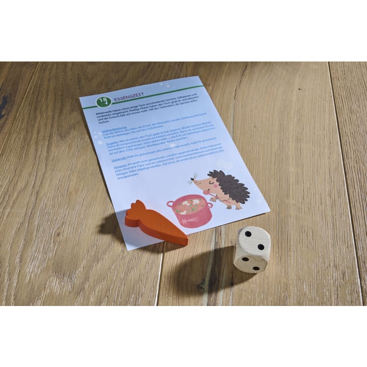 Eine Spielanleitung mit deutschem Text, die Illustration eines Igels neben einem Topf und zwei Spielfiguren - eine hölzerne Karotte und ein weißer Würfel mit schwarzen Punkten - liegen auf einer Holzunterlage.