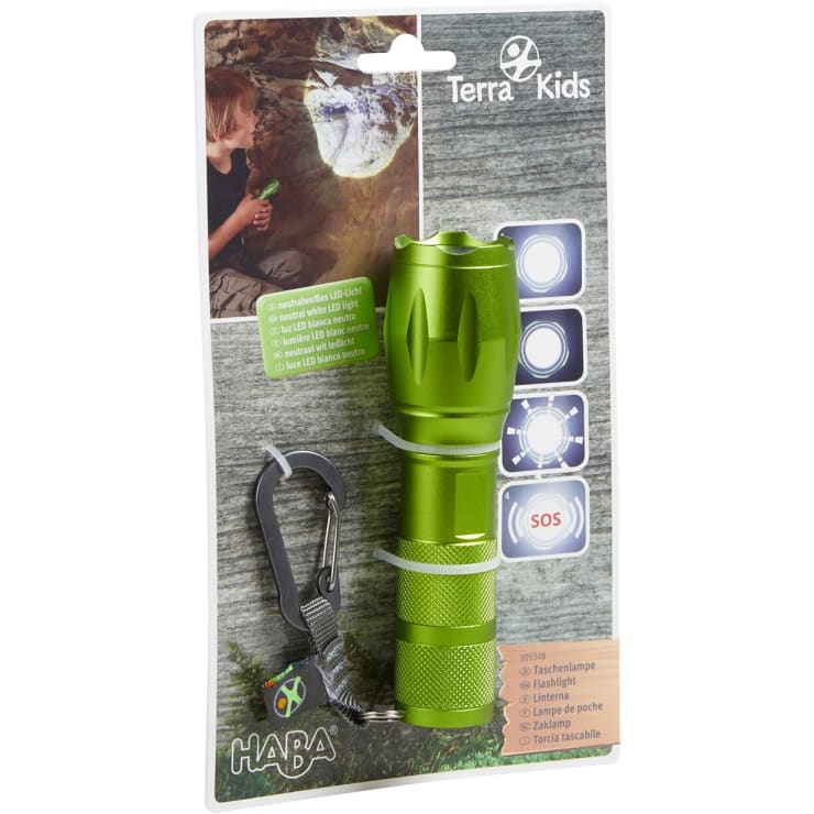 Eine grüne Taschenlampe mit einem Karabinerhaken ist auf einer Karte mit der Aufschrift Terra Kids verpackt. Auf der Verpackung sind LED-Lichtmodi abgebildet, darunter weiß, neutral und SOS. Ein Foto zeigt ein Kind, das die Taschenlampe in einer Höhle benutzt.