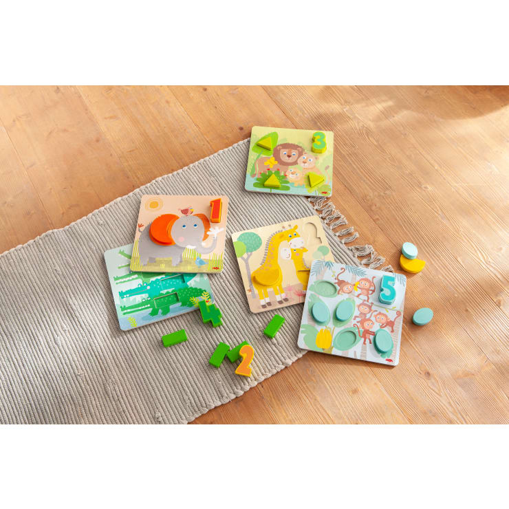 Vier bunte Holzpuzzlebretter mit Zahlen und passenden 3D-Teilen liegen auf einem beigen Teppich auf einem Holzboden. Die Zahlen 1, 2, 3 und 5 sind auf den Puzzlebrettern zu sehen.