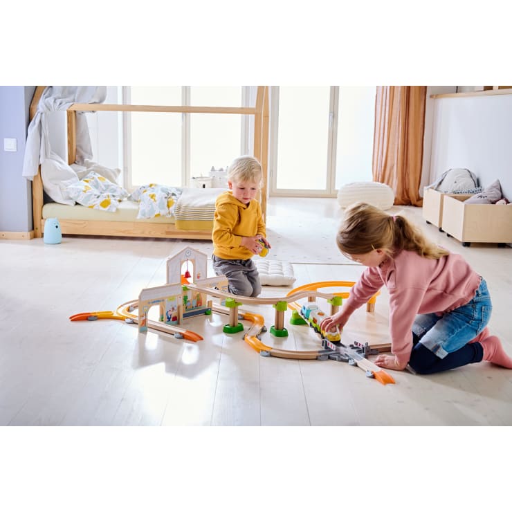 Zwei kleine Kinder spielen mit einer Holzspielzeugeisenbahn auf dem Boden eines hellen, aufgeräumten Schlafzimmers mit einem Bett und großen Fenstern im Hintergrund. Die Kinder konzentrieren sich auf die Eisenbahnschienen und das Zubehör.