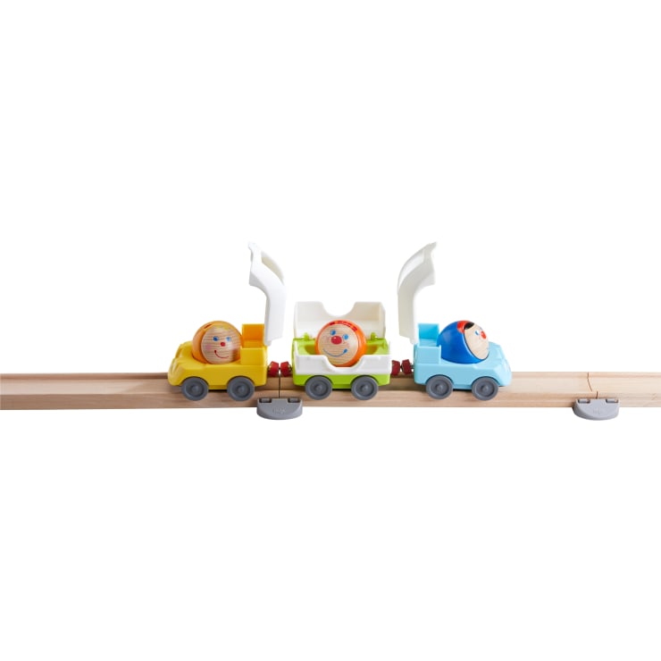 Ein hölzerner Spielzeugeisenbahnzug mit drei bunten Waggons, die jeweils eine runde Holzfigur mit einem bemalten Gesicht tragen, steht auf einer Holzschiene mit weißem Hintergrund.