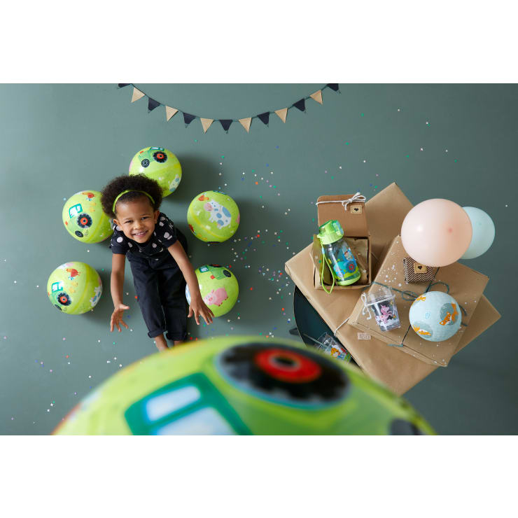 Ein kleines Kind in einem schwarzen Overall sitzt auf dem Boden, umgeben von bunten Luftballons und Konfetti, daneben ein Stapel eingepackter Geschenke und pastellfarbener Luftballons. An der Wand darüber hängt ein dekoratives Wimpel-Banner.
