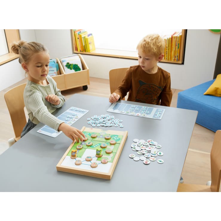 Zwei Kinder sitzen an einem Tisch und spielen ein Brettspiel mit bunten, kreisförmigen Figuren. Im Hintergrund befinden sich Regale mit Büchern und ein blauer Sitz. Das Mädchen streckt die Hand aus, um eine Figur auf dem Spielbrett zu bewegen.