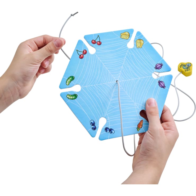 Zwei Hände halten eine sechseckige blaue Karte mit einem weißen Spinnennetzmuster, Lebensmittelillustrationen an jeder Ecke und einer durch Schlitze geführten Schnur. Ein gelber Würfel mit Bildern ist ebenfalls zu sehen.