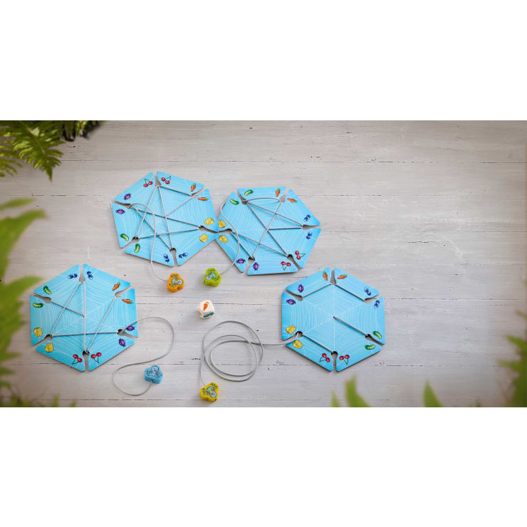 Drei blaue sechseckige Spielbretter mit bunten Fußabdrücken sind durch graue Schnüre miteinander verbunden. Vier kleine Tierfiguren und zwei Würfel stehen neben den Brettern auf einer hellen Holzfläche, die von grünen Blättern eingerahmt wird.