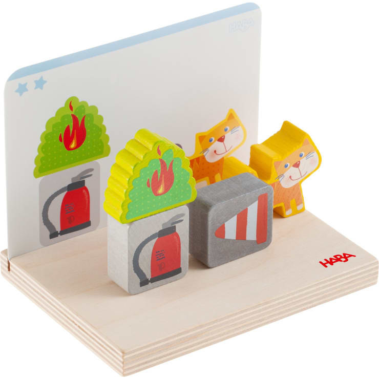 Ein Holzspielzeugset mit bemalten Blöcken, darunter zwei Bäume mit Flammen, ein grauer Block mit einem Barrieremuster, zwei orangefarbene Katzen und ein weißer Hintergrund mit blauen Sternen. Der Sockel ist aus hellem Holz und mit der Aufschrift "HABA" versehen.