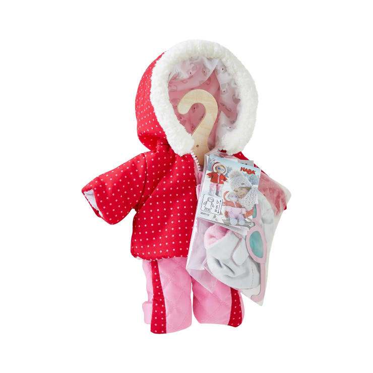 Eine hölzerne Schaufensterpuppe ist mit einer rot-weiß gepunkteten Kapuzenjacke mit weißem Pelzbesatz und einer rosa Hose bekleidet. Die Schaufensterpuppe hält eine durchsichtige Tasche mit Puppenzubehör, einschließlich Kleidungsstücken, in der Hand.