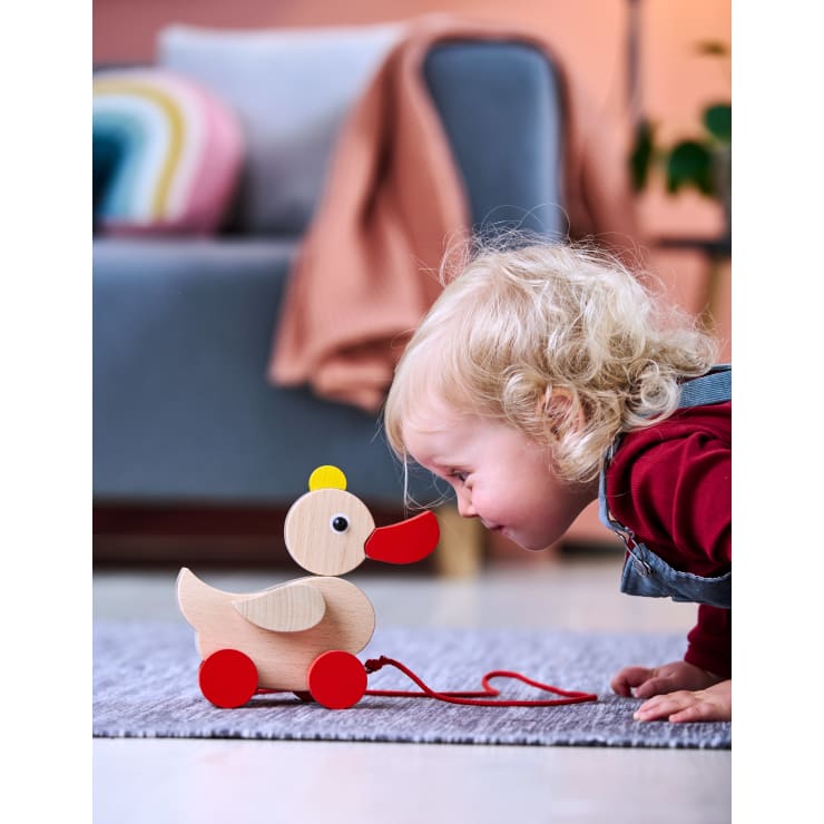 Ein kleines Kind mit blondem Haar lehnt nach vorne auf einem grauen Teppich, dicht vor einem hölzernen Nachziehspielzeug in Form einer Ente mit roten Rädern. Im Hintergrund ist ein Sofa mit einer Decke und einem Kissen zu sehen.