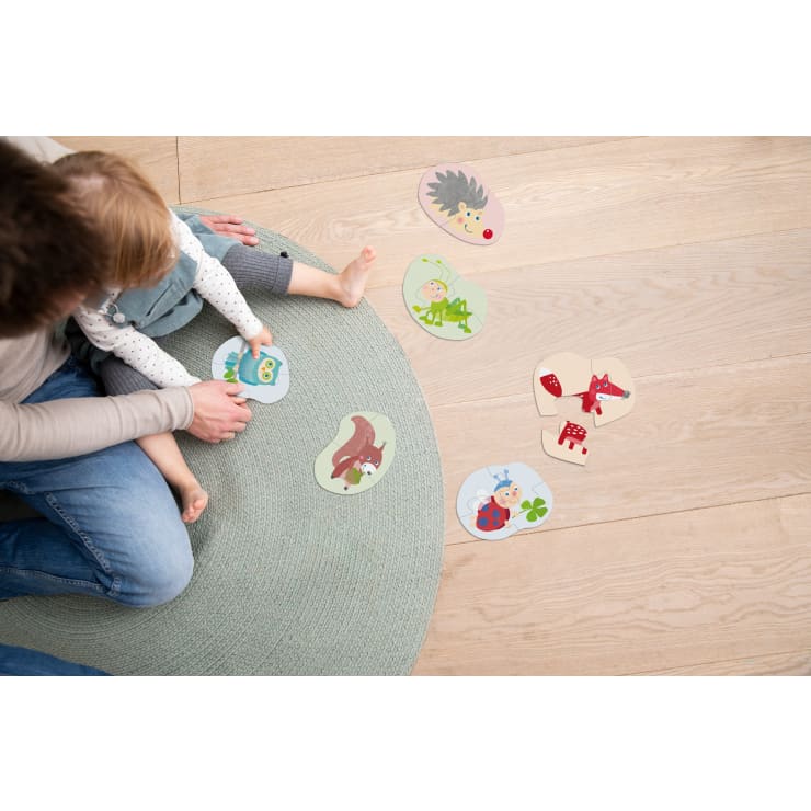 Ein Erwachsener und ein Kleinkind sitzen auf einem runden, hellblauen Teppich und setzen auf einem Holzboden Puzzleteile mit Tiermotiven zusammen. Mehrere fertige und nicht zusammengesetzte Puzzleteile liegen in der Nähe verstreut.