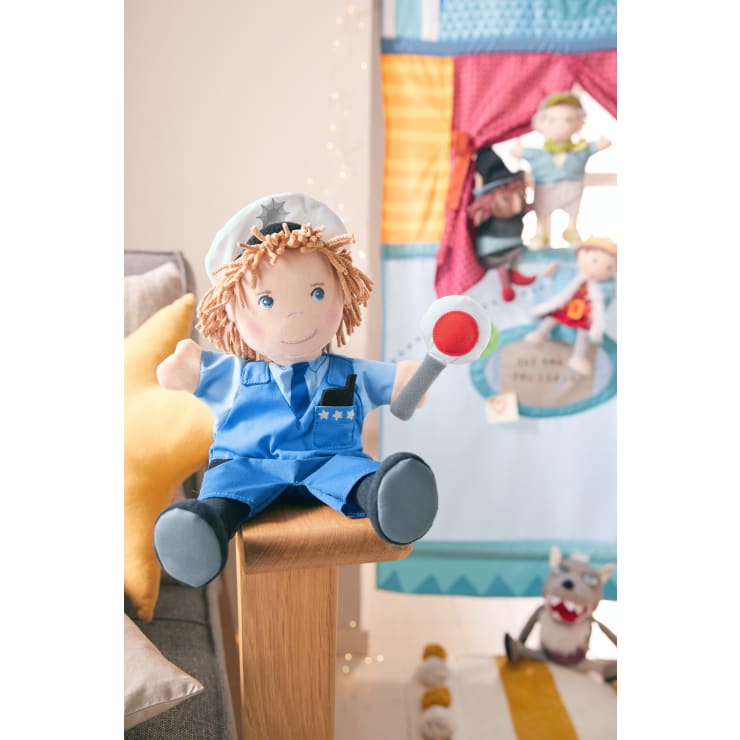 Eine als Polizist verkleidete Plüschpuppe sitzt auf der Armlehne eines Holzstuhls und hält einen Spielzeugschlagstock. Im Hintergrund ist ein buntes Puppentheater mit anderen Plüschpuppen und Spielzeug zu sehen.