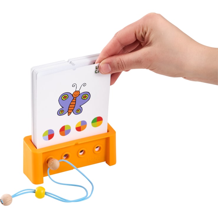 Eine Hand steckt eine Karte mit einer bunten Schmetterlingsillustration und farbigen Kreisen in einen orangefarbenen Kartenhalter, an dem blaue Gummibänder mit Holzperlen befestigt sind.