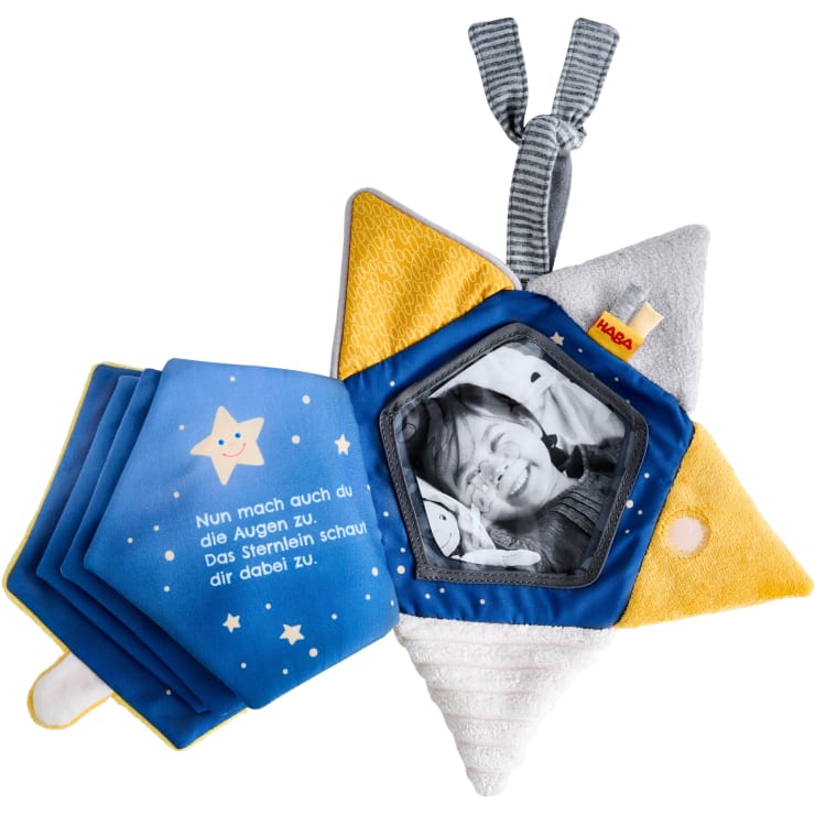 Ein weiches, sternförmiges Babyspielzeug mit strukturierten gelben, weißen und grauen Punkten, ein angebrachter Fotohalter aus Stoff, der ein Baby zeigt, und ein Stoffbuch mit einer deutschen Botschaft und Sternenillustrationen.