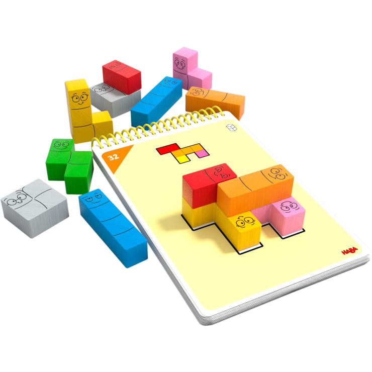 Bunte Holzklötze mit Gesichtern sind um und auf einem spiralgebundenen Puzzlebuch angeordnet. Die aufgeschlagene Seite zeigt eine Aufgabe mit einem Blockmuster, das mit den Blöcken nachgebaut werden soll.